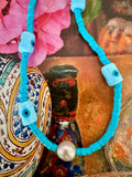 A turquoise beaded necklace with square evil eye charms and a central white freshwater pearl.