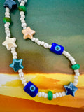 A beaded necklace featuring white round beads, blue evil eye square beads, teal and blue star-shaped beads, and small green cylindrical beads.
