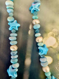 A close-up of a beaded strand featuring light blue and white starfish beads alternating with smooth, polished pebble beads in shades of light blue, white.