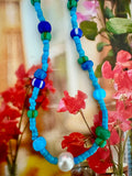 A beaded necklace featuring turquoise blue beads, dark blue and green accent beads, and a single white freshwater pearl.