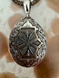 An oval silver locket pendant with an ornate engraved cross design, hanging from a silver bail on a beaded necklace, set against a textured beige fabric background.