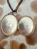 An open silver locket pendant necklace on a dark beaded chain, displayed on a textured beige and brown patterned fabric background.