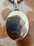 A close-up of a polished oval silver locket pendant on a chain. The silver surface has a small maker's mark stamped near the bottom.